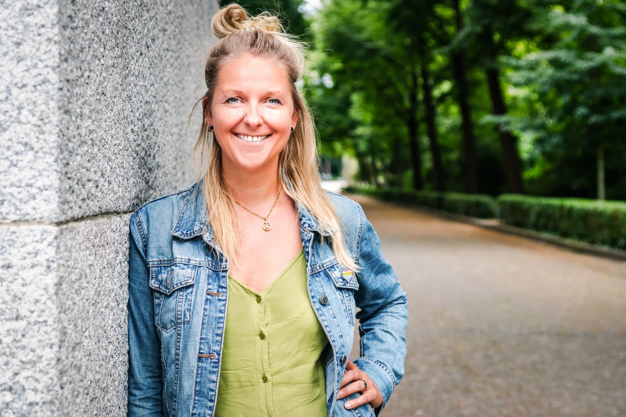 Jenny Heyden in Jeansjacke lehnt lächelnd an einer Steinmauer, dahinter eine grüne Allee